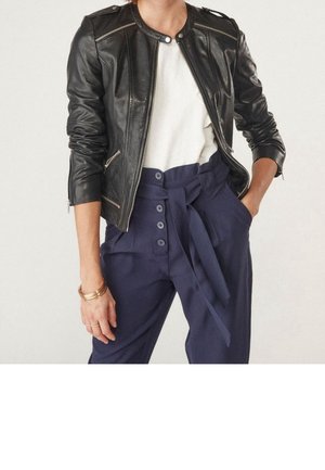 Woman wearing a black leather jacket over a white shirt, navy high-waisted pants with buttons and a tied belt, with hands in pockets.