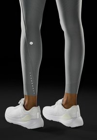 Szare legginsy z teksturowanej tkaniny, z logo na udzie i kropkowymi akcentami w okolicy kostki, zestawione z białymi butami sportowymi.