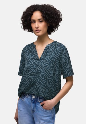 Femme aux cheveux courts et bouclés portant un chemisier à rayures zébrées vert foncé et noir et un jean bleu clair, avec une main dans la poche.