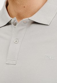 Jasnoszara koszulka polo z teksturowanej tkaniny, z kołnierzykiem i zapięciem na trzy guziki. Posiada subtelne haftowane logo na piersi.
