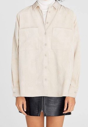 Chemise beige à boutons avec deux poches poitrine portée sur un col roulé blanc, associée à une mini-jupe noire texturée avec une fermeture éclair frontale.