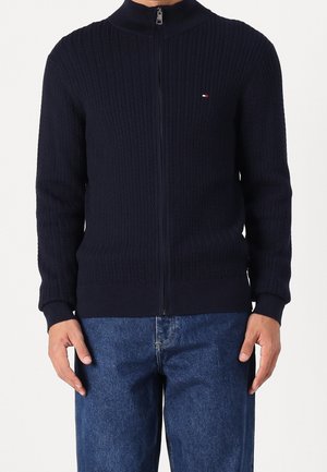 Man draagt een marineblauwe, gebreide trui met ritssluiting en een klein logo op de borst, gecombineerd met blauwe spijkerbroek tegen een effen achtergrond.