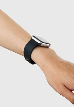 Smartwatch med sort rem, båret på venstre håndled mod en ensfarvet lys grå baggrund.