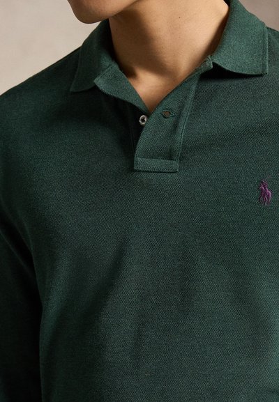 Muškarac nosi tamnozeleno polo majicu s ljubičastim izvezenim logotipom konja na prsima i ovratnikom sa jednim zakopčanim gumbom