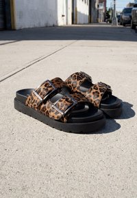 Paire de sandales à imprimé léopard avec semelles épaisses noires et boucles argentées sur un trottoir en béton urbain au soleil.