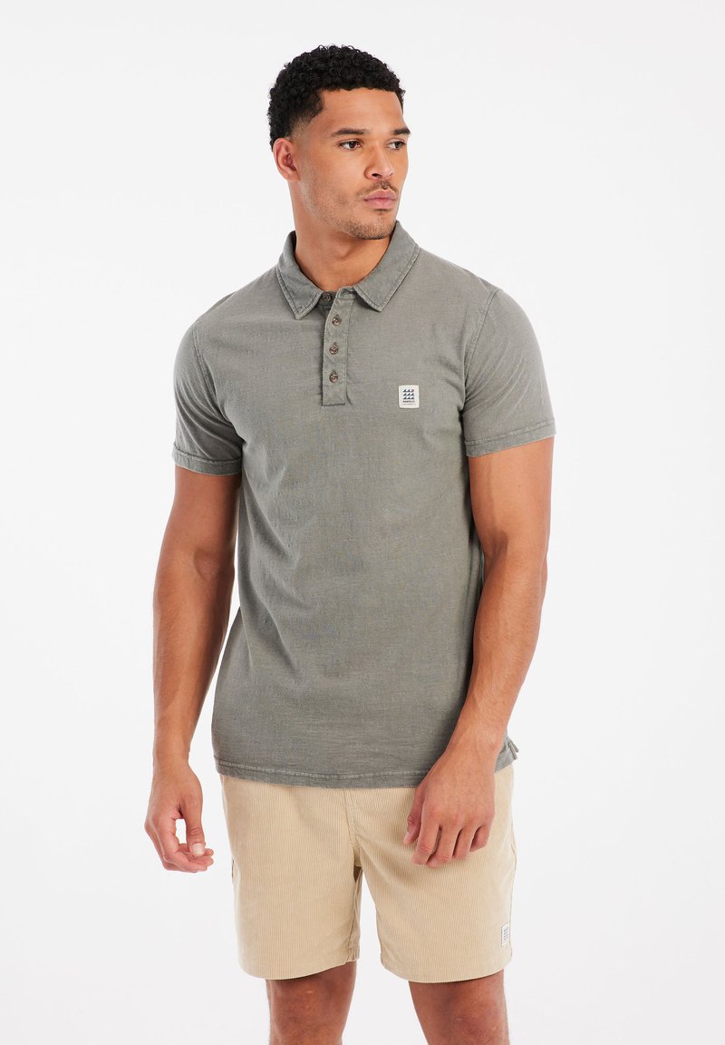 Olivengroen polo met een kraag, vijfknops sluitsysteem en een klein logo-embleem. Gecombineerd met lichtbeige gestreepte shorts.