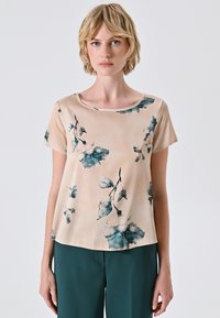 Blusa a stampa floreale in beige chiaro con accentuazioni blu-verdi, maniche corte e scollo rotondo. Tessuto leggero e liscio con una vestibilità rilassata.