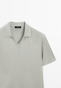 Hellgraues Polohemd mit kurzen Ärmeln, weichem Kragen und V-Ausschnitt, flach auf weißem Hintergrund liegend.