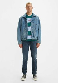 Giacca di jeans azzurro chiaro sopra una camicia a righe verdi, blu navy e bianche, abbinata a jeans scuri e sneaker alte.