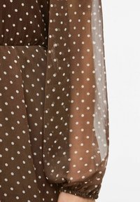 Brun prikkete kjole med gjennomsiktige lange ermer, hvite prikker, elastiske mansjetter og et flytende design. Strukturert stoff gir visuell dybde.