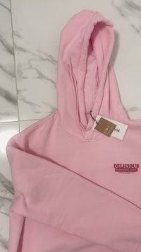 Hoodie rosa claro feito de tecido suave, apresentando um bolso canguru e um logotipo impresso "FRUTO REFRESCANTE E DELICIOSO" em vermelho escuro.