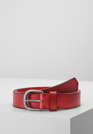 Punainen nahkavyö hopeanvärisellä suorakaiteen muotoisella soljella, kierrettynä valkoisella pinnalla vaaleanharmaata taustaa vasten.