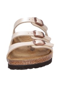 Vit sandal med tre remmar och bronsfärgad spännen, korkfotbädd och texturerad svart sula. Platt design som passar för avslappnad användning.