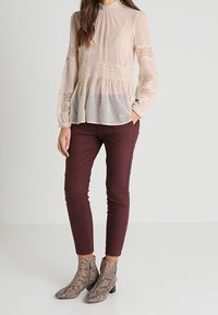 Helle beige Bluse mit langen Ärmeln, strukturierten gepunkteten Motiven, Rüschenausschnitt und Peplum-Saum, kombiniert mit burgunderroten, eng anliegenden Hosen und ankle boots mit Schlangenlederoptik.