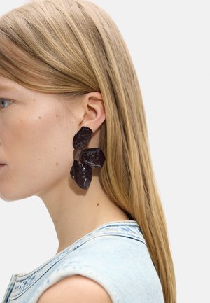 Blondynka w jasnoniebieskim bezrękawniku z dużymi, abstrakcyjnymi, ciemnobrązowymi kolczykami w kształcie kwiatów na lewym uchu.