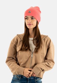 Giovane donna con un berretto corallo, una felpa beige con zip e jeans blu, che guarda leggermente di lato con le mani vicino al punto vita.