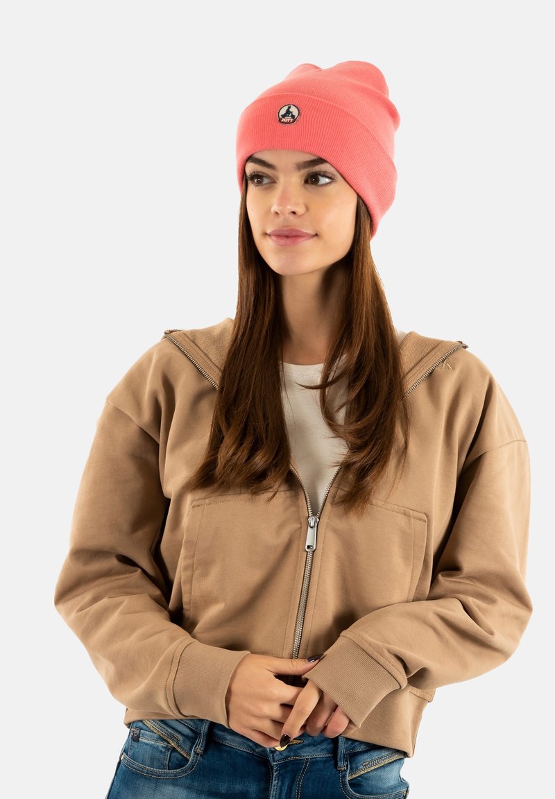 Giovane donna con un berretto corallo, una felpa beige con zip e jeans blu, che guarda leggermente di lato con le mani vicino al punto vita.