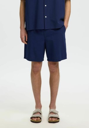 Personne portant un short bleu marine et une chemise à manches courtes assortie, sandales beiges, debout avec une main dans la poche sur fond blanc.