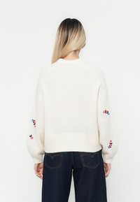 Weißer Strickpullover mit geripptem Saum und langen Ärmeln, die mit floralem Stickerei in Pink und Blau versehen sind.