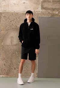 Svart faux fur zip-up hettejakke med frontlommer, iført over en hvit skjorte. Kombinert med svarte shorts og hvite sneakers, står mot en teksturert vegg.