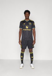 Μαύρη αθλητική στολή ποδοσφαίρου με κίτρινες λεπτομέρειες, συμπεριλαμβανομένου του γιακά και των λογότυπων. Συνδυάζεται με αντίστοιχα σορτς και κάλτσες, καθώς και με λευκά παπούτσια με σ studs.