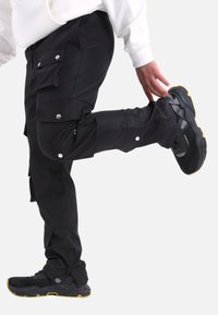 Schwarze Cargo-Hosen mit mehreren Taschen, silbernen Druckknöpfen und Reißverschlüssen an den Säumen, kombiniert mit robusten schwarzen Sneakers mit gelben Akzenten.