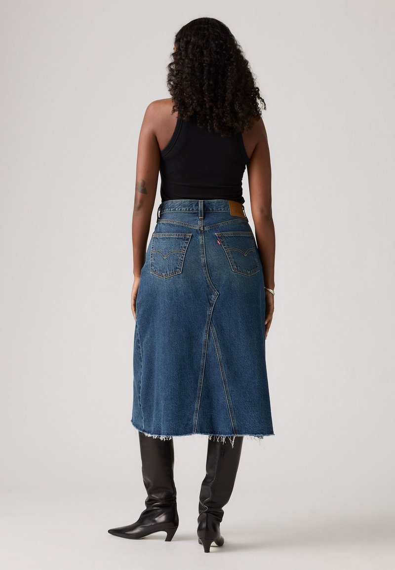 Jupe midi en denim bleu foncé avec une finition brute, des poches arrière et une taille haute. Associée à des bottes noires montantes.