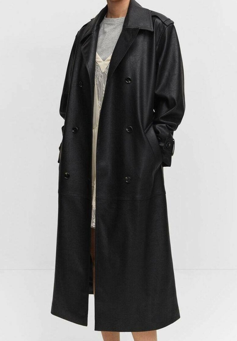 Personne portant un long manteau en cuir noir croisé avec de larges revers et les mains dans les poches, par-dessus un haut gris et une robe en dentelle.