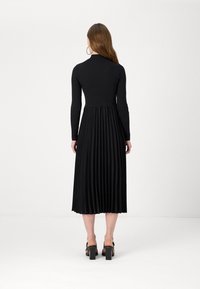 Mujer con cabello largo castaño llevando grandes pendientes de aro, un vestido midi plisado negro de manga larga y sandalias de tacón alto negras, de espaldas.