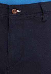 Gros plan de la ceinture d’un pantalon bleu marine foncé avec un bouton marron et une passants de ceinture sur un fond en tissu bleu.