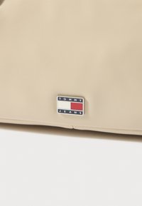 Τσάντα από συνθετικό δέρμα σε μπεζ χρώμα με μεταλλική ταμπέλα λογότυπου "TOMMY JEANS" σε ναυτικό μπλε, λευκό και κόκκινο. Λείο υλικό με ελάχιστες λεπτομέρειες.