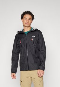 Veste noire imperméable avec une capuche grise, poches zippées sur la poitrine et poignets ajustables. Présente des tirettes de fermeture éclair et un logo rouges contrastants.