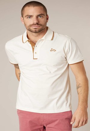 Homme aux cheveux courts et barbe portant un polo blanc avec bordure marron et un short rose, debout devant un fond beige uni.