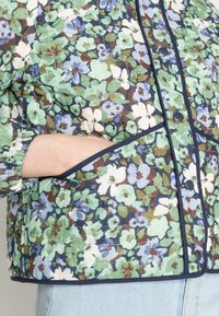 Gewatteerd bloemenjack met groene, blauwe en witte bloemen op een marineblauwe achtergrond, voorzien van een ronde kraag en zijzakken.