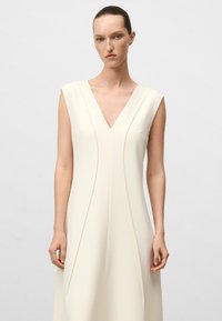 Ärmelloses V-Ausschnitt-Kleid aus weichem Elfenbein-Stoff, mit vertikalen Nahtdetails und einem glatten, minimalistischen Design.