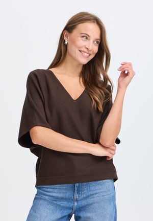 Jeune femme aux longs cheveux bruns sourit en regardant vers la droite, portant un haut marron foncé à col en V et un jean bleu clair sur un fond uni.