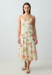 Robe midi florale avec bretelles fines, présentant un fond crème clair orné de pivoines roses et de feuilles vertes. Tissu lisse et fluide.
