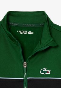 Zelena zip-up jakna iz teksturiranega materiala, z črnim akcentom in svetlo modro črto. Vključuje Lacoste logo na prsih.