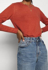Maglione a coste di colore ruggine con maniche lunghe, abbinato a jeans in denim leggeri a vita alta di colore blu con un classico design a cinque tasche.