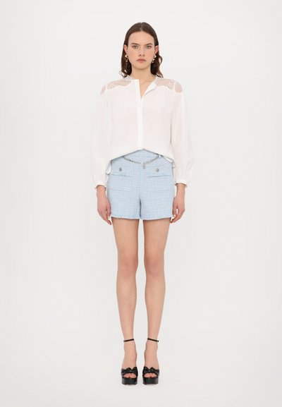 Blusa blanca con acentos de encaje en los hombros, combinada con pantalones cortos de tweed azul claro con dobladillo deshilachado y bolsillos frontales, llevada con tacones negros.