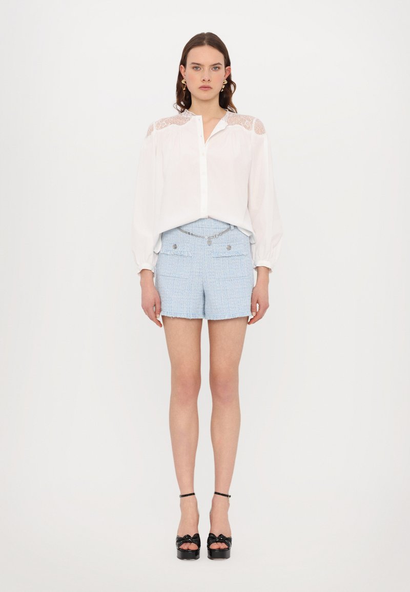 Blusa blanca con acentos de encaje en los hombros, combinada con pantalones cortos de tweed azul claro con dobladillo deshilachado y bolsillos frontales, llevada con tacones negros.