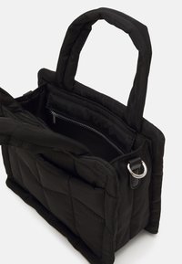 Sac cabas noir matelassé avec anses rembourrées, ouverture supérieure dévoilant une poche intérieure zippée et un anneau métallique argenté sur le détail latéral.