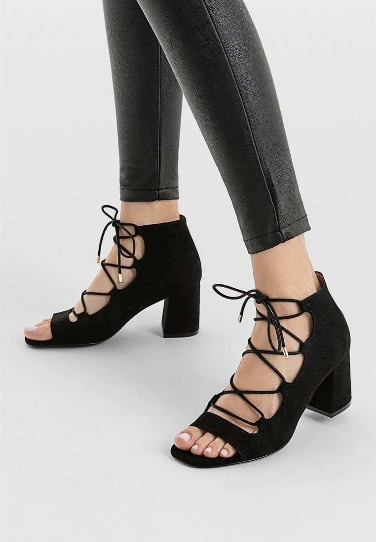 Pieds dans des chaussures noires à talons blocs, lacées et à bout ouvert, associées à un pantalon noir ajusté sur un fond gris clair.