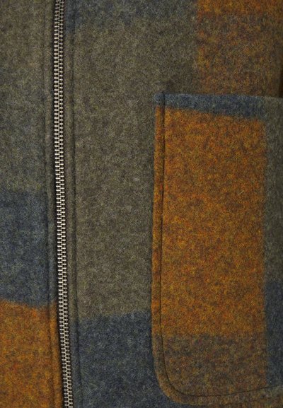 Wool blend jacket featuring a zip closure, green and brown plaid pattern, with a patch pocket in orange and brown shades. Textured surface detail.