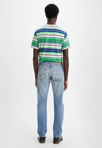 Persona vistiendo una camiseta a rayas verde, azul y blanca con jeans azul claro y zapatos negros, de pie de espaldas sobre un fondo liso.