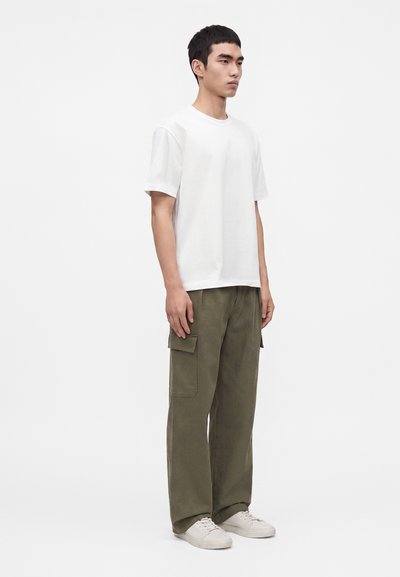 Jeune homme debout, face à droite, portant un t-shirt blanc uni, un pantalon cargo vert olive et des baskets blanches sur fond blanc.
