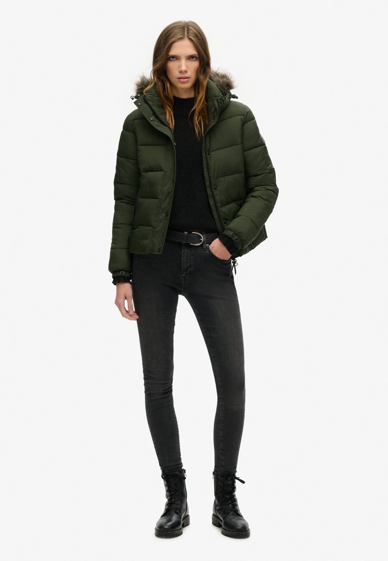 Giacca a vento verde scuro con cappuccio foderato in pelliccia, zip frontale e polsini elasticizzati, abbinata a un maglione nero e jeans skinny grigi.