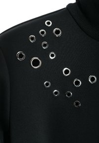 Sudadera negra de cuello alto con ojales plateados de diferentes tamaños dispuestos en un hombro, creando un patrón texturizado.