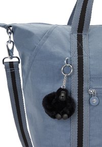 Modrá látková taška se strukturovaným tvarem. Obsahuje černě pruhované popruhy a chlupatý klíčenkový přívěsek gorily. Má také kapsu na zip a kovové detaily.