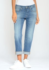 Mujer vistiendo jeans claros con dobladillos y zapatillas blancas, de pie contra un fondo liso.